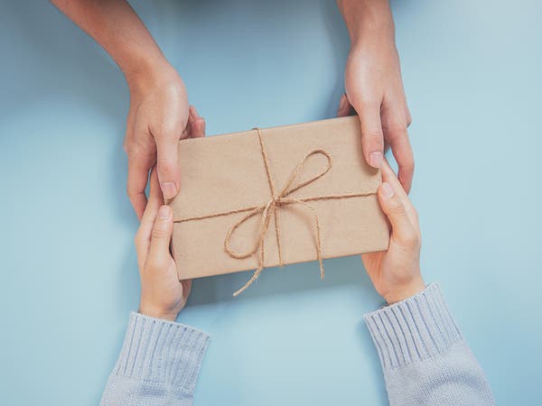 Des mains d'enfant en train de remettre un cadeau emballé dans du papier craft beige à des mains d'adulte, sur un fond bleu pale
